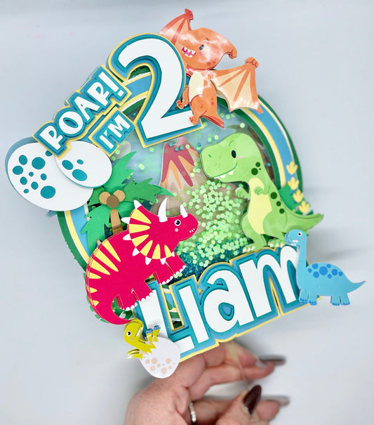A colorful dinosaur-themed birthday cake topper with the text 'I'm 2!' and various dinosaur illustrations, held by a hand with artificial nails.